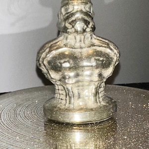 May include: A silver-colored bust of a Stormtrooper, a character from the Star Wars franchise. The bust is detailed with the helmet and chest armor. It is sitting on a round, glittery, silver-colored surface.