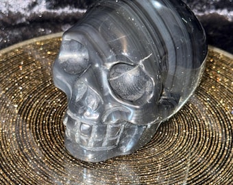 May include: A polished, dark gray skull figurine with a striped pattern. The skull has detailed features, including eye sockets, a nose cavity, and teeth. The figurine is displayed on a gold, glittery surface.