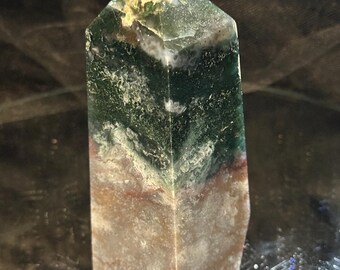 Puede incluir: Un obelisco de piedra preciosa rectangular y puntiagudo. La piedra presenta capas de tonos verdes, blancos y marrones. El obelisco está de pie, mostrando sus patrones y texturas naturales. La piedra preciosa está pulida y refleja la luz.