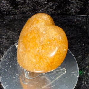 May include: A golden-orange heart-shaped crystal. The polished stone has a textured surface and sits on a clear acrylic stand. The crystal is displayed against a dark, textured background.