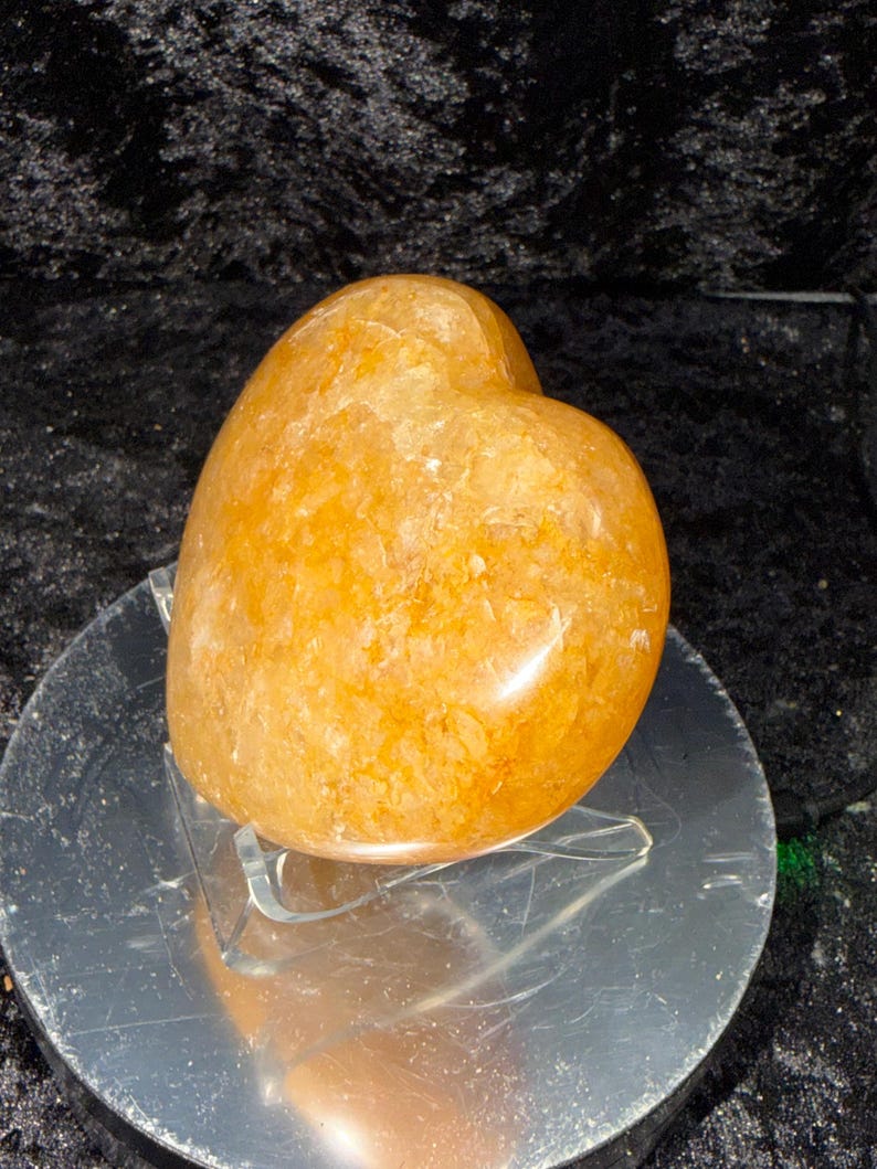 May include: A golden-orange heart-shaped crystal. The polished stone has a textured surface and sits on a clear acrylic stand. The crystal is displayed against a dark, textured background.