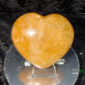 May include: A golden-orange heart-shaped crystal, polished to a smooth finish. The gemstone sits on a clear acrylic stand, displayed against a dark background. The heart's color ranges from light to deep orange.