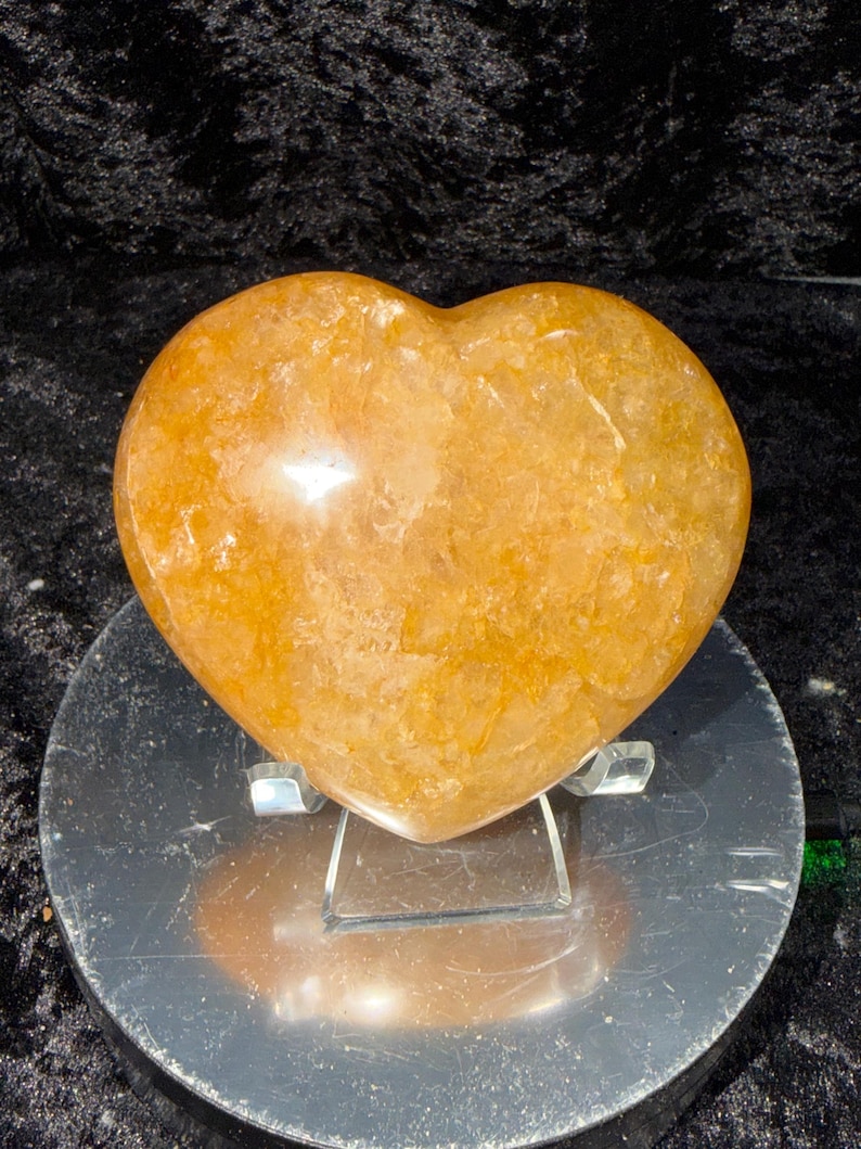 May include: A golden-orange heart-shaped crystal, polished to a smooth finish. The gemstone sits on a clear acrylic stand, displayed against a dark background. The heart's color ranges from light to deep orange.