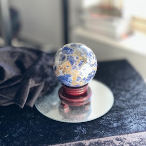 May include: A blue and gold sphere with a marbled pattern sits on a wooden stand. The sphere is on a mirrored surface.