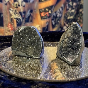 May include: Two gray, crystal-like stones with a sparkly, glittery texture, sitting on a round, silver, glittery plate.