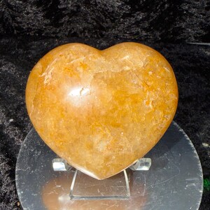 May include: A heart-shaped, polished stone in shades of orange and yellow. The stone has a smooth, glossy surface with natural variations in color and texture. It is displayed on a clear acrylic stand, showcasing its shape and color.