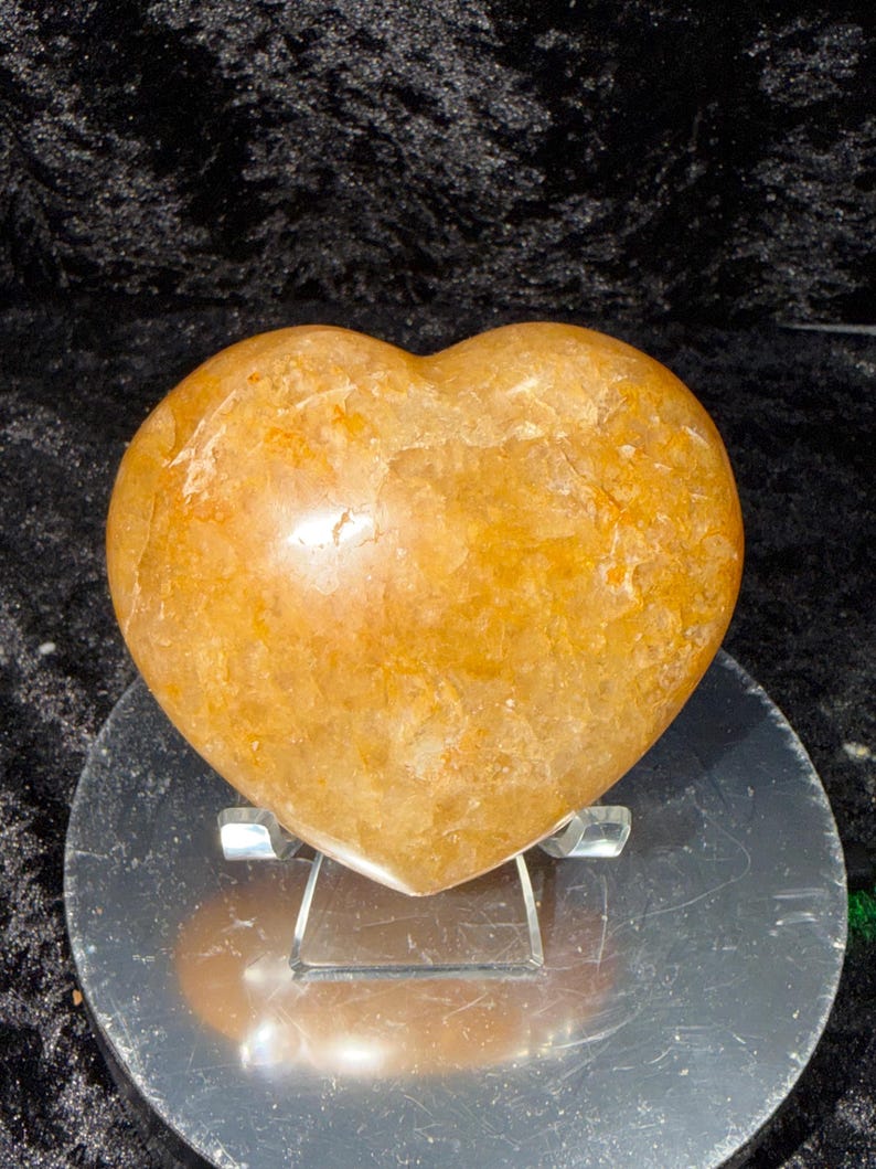 May include: A heart-shaped, polished stone in shades of orange and yellow. The stone has a smooth, glossy surface with natural variations in color and texture. It is displayed on a clear acrylic stand, showcasing its shape and color.