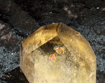 May include: A faceted, translucent, golden-yellow crystal with a geometric shape. The crystal has internal iridescent inclusions and sits against a dark, textured background. The crystal's facets catch the light, creating highlights.
