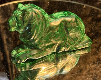 May include: A carved malachite figurine in the shape of a reclining lion. The stone features deep green and light green concentric banding patterns throughout its surface, with a smooth, polished finish that reflects light across its curved form.
