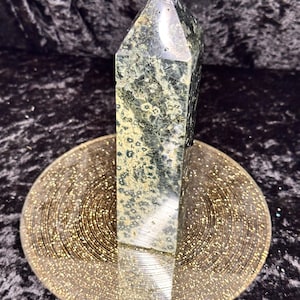 May include: A green and black obelisk shaped crystal with a white top, sitting on a gold glitter-covered plate.