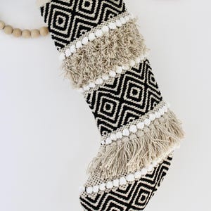 May include: A Christmas stocking with a geometric black and white pattern. The stocking features beige fringe and white pom-poms. A beige fabric is used for the top and the hanging loop. The stocking is decorated with a diamond pattern.