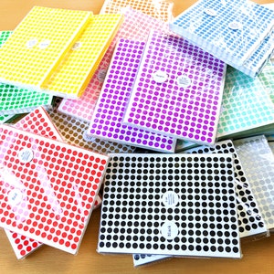 Könnte beinhalten: Eine Sammlung von bunten Punkt-Stickern in verschiedenen Farben, darunter Rot, Lila, Schwarz und Blau. Die Sticker sind in Stapeln angeordnet und mit dem Farbnamen beschriftet.