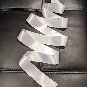 May include: A length of white satin ribbon, arranged in a flowing, serpentine pattern. The ribbon's smooth texture reflects light, creating highlights and shadows. The ribbon is against a black, textured background.
