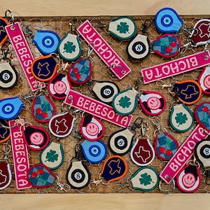 May include: A collection of colorful keychains with various designs, including a blue eye, a red heart, a green clover, a yellow smiley face, and a red and white striped design. The keychains are made of fabric and have a metal ring for attaching to keys. The keychains are arranged on a wooden surface with a wire mesh background.