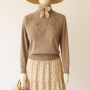 May include: A vintage-style outfit on a mannequin. The ensemble includes a tan sweater with floral embellishments, a floral print skirt, a straw hat, and a matching scarf. A small woven basket with flowers completes the look.