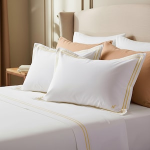 May include: White bed linens with gold trim and accent pillows. The pillows and sheets have a gold border. The bed is made with white sheets and pillowcases. The pillows are arranged on the bed with a neutral-colored headboard in the background.