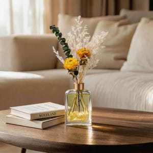May include: A clear glass rectangular bottle with a gold cap containing yellow liquid and small yellow stones at the base, labeled with the text Netbors. A bouquet of dried yellow flowers, white filler plants, and green eucalyptus stems sits inside.