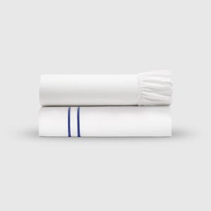 May include: Two folded white bed sheets. The bottom sheet has two vertical blue stripes. The top sheet is rolled and has a ruffled edge. The sheets are stacked on top of each other against a white background.