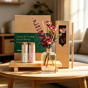 May include: A reed diffuser fragrance set featuring a clear glass bottle with pink crystals, a gold cap, and dried red roses and eucalyptus stems. Includes two small glass vials with cork stoppers containing white and pink crystals, and a brown box.