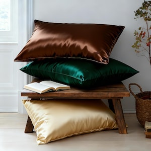 May include: Three satin pillowcases in shades of brown, emerald green, and gold are stacked on a rustic wooden bench. An open book rests on the bench between the green and gold pillowcases. The pillows have a shiny, smooth texture.