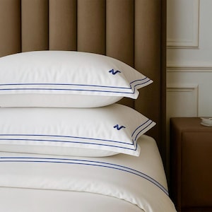 May include: White bed linens with blue trim and embroidered details. The image shows a stack of pillows and a duvet cover, all featuring a clean, crisp design. The blue accents provide a classic, elegant touch to the bedding.