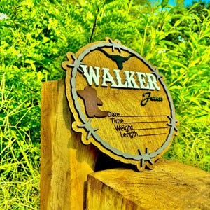 May include: A wooden sign with a rustic design featuring a barbed wire border and a silhouette of a longhorn steer. The sign is personalized with the name "WALKER" and includes spaces to record the date, time, weight, and length.