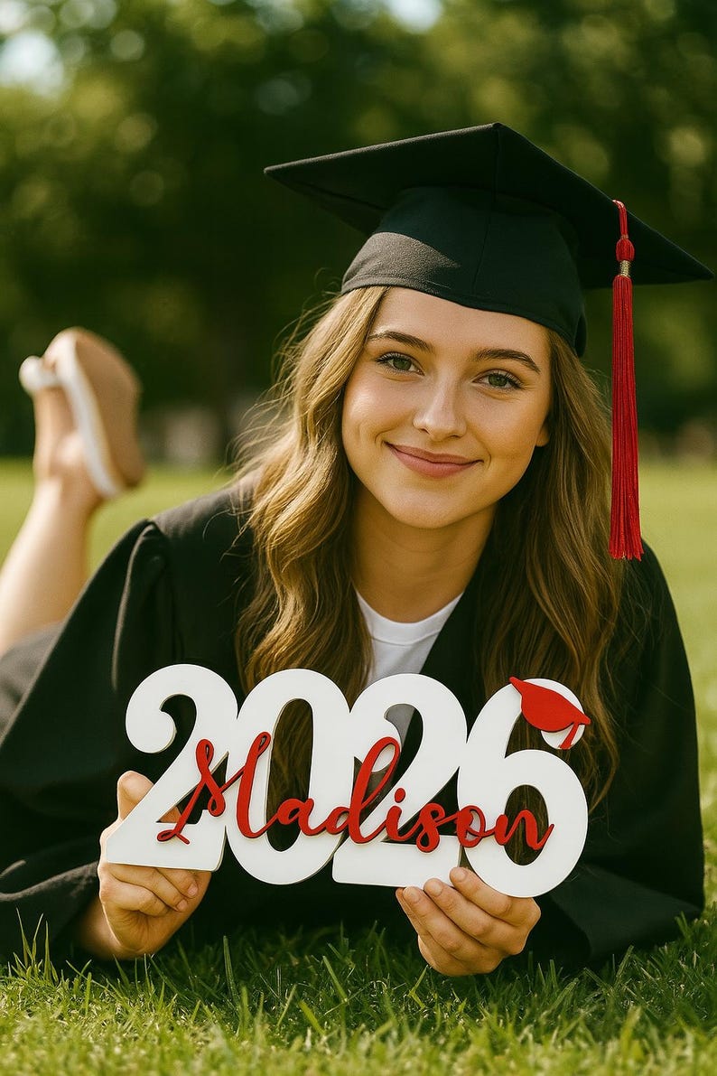Peut inclure: Photo de remise de dipl&ocirc;me avec une personne portant une toque et une toge noires, tenant une pancarte blanche avec l'ann&eacute;e 2026 et le nom "Madison" en rouge. La pancarte a un petit chapeau de remise de dipl&ocirc;me.
