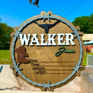May include: A wooden round sign with a barbed wire border, a brown cowboy boot, a black and white steer head, and the name "WALKER James" in white letters. The sign has spaces to fill in the date, time, weight, and length.
