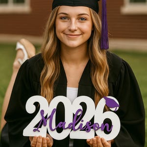 Peut inclure: Une personne portant une robe et un chapeau de graduation noirs avec un gland violet, tenant une pancarte en bois blanche avec "2026 Madison" en script violet. L'arri&egrave;re-plan comprend de l'herbe verte et un b&acirc;timent en briques.