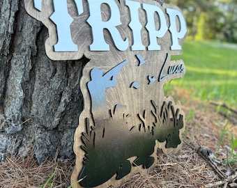 May include: A wooden sign with the name "TRIPP" in white letters on a brown background. The sign features a silhouette of a person hunting ducks with the text "Lucas" below the silhouette.