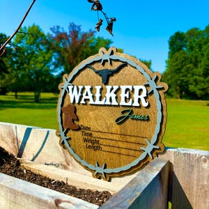 May include: A wooden sign with a rustic brown finish and a blue and green barbed wire design. The sign says "WALKER James" and has spaces to fill in the date, time, weight, and length.