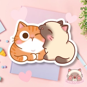 以下が含まれることがあります： 2匹の漫画の猫が描かれたステッカー。1匹はオレンジ色の縞模様、もう1匹は茶色のアクセントが付いたクリーム色です。彼らは互いに近く、1匹の猫がもう1匹を寄り添っています。ピンクのハートと「TOEBEAN TOWN」の文字も表示されています。
