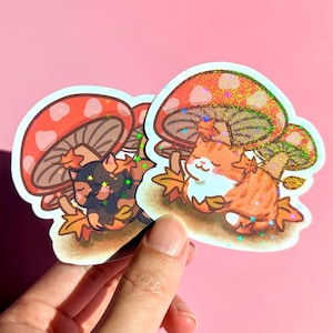 May include: Two stickers depicting cartoon cats under mushrooms. One cat is black with gold stars, the other is orange and white. The mushrooms have red caps with white spots and glittery details. Autumn leaves surround the cats.