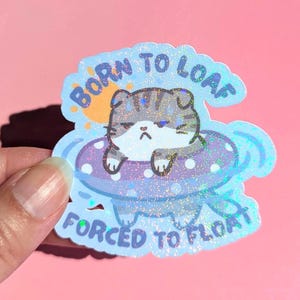 May include: A sticker featuring a grumpy cartoon cat in a polka-dot inner tube. The sticker has the text "BORN TO LOAF FORCED TO FLOAT" in a blue, glittery font. A small sun is in the upper left corner.