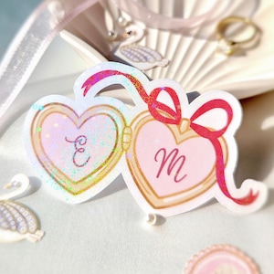 May include: A heart-shaped sticker with a gold border, featuring the letters "E" and "M" inside. The sticker has a pink background with a glittery effect and a red ribbon. The sticker is a romantic item.
