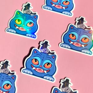 May include: A collection of stickers featuring a blue cat with orange eyes, a wide grin, and a small bird wearing a hat. The stickers have a holographic effect and are set against a pink background.