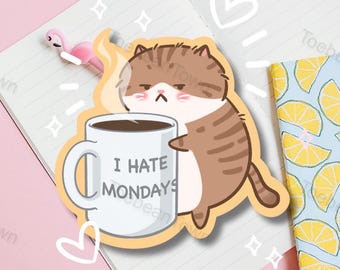 May include: A cartoon cat holding a white coffee mug with the text "I hate Mondays". The cat is brown and white with a grumpy expression. The mug is filled with brown coffee and has steam coming from it.