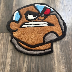 May include: A brown, black, and gray rug shaped like a cartoon character's head. The rug features a brown face with white teeth, a gray metallic section, and blue and red accents. The rug is placed on a wooden floor.