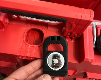 Puede incluir: Un soporte negro con un AirTag de Apple adjunto, sobre una caja de herramientas roja. El AirTag es plateado con el logotipo de Apple. El soporte tiene un lazo para sujetarlo.