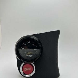 May include: A black automotive gauge with a round display showing air/fuel ratio, and a red "ENGINE START" button. The gauge is mounted on a black textured panel. The gauge face has white markings and the brand name "AEM".