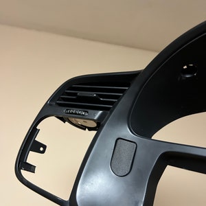 May include: Black plastic dashboard panel for a car. The panel has a vent, a small button, and a rectangular cutout.