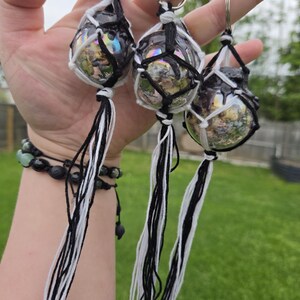 May include: Three glass orb keychains, each encased in a macrame net with black and white cord tassels. The orbs have a reflective, iridescent quality, and are suspended from silver keyrings. The background is a blurred green lawn.