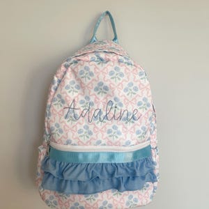 May include: A white backpack with a pink and blue floral pattern. The backpack has a blue ruffle trim and the name "Adaline" embroidered on the front.