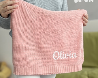 May include: A pink knitted baby blanket with the name "Olivia" embroidered in white.