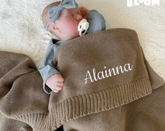May include: A brown knitted baby blanket with the name "Alaina" embroidered in white. The blanket is folded and a baby doll is lying on top of it.