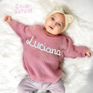 May include: A pink knitted sweater with the name "Luciana" embroidered in white on the front. The sweater is worn by a baby wearing a white headband with a bow.