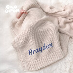 Op de afbeelding: Een beige gebreide babydeken met de naam "Brayden" in blauw geborduurd. De deken is opgevouwen, met een babyhandje zichtbaar. De deken heeft een geribbelde rand en de tekst "Coco Bloom" in de linkerbovenhoek.