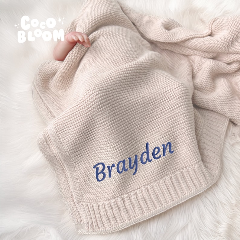 Op de afbeelding: Een beige gebreide babydeken met de naam "Brayden" in blauw geborduurd. De deken is opgevouwen, met een babyhandje zichtbaar. De deken heeft een geribbelde rand en de tekst "Coco Bloom" in de linkerbovenhoek.