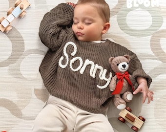 May include: A brown knitted sweater with the name "Sonny" embroidered in white on the front. A brown teddy bear with a red bow is held by the child wearing the sweater.