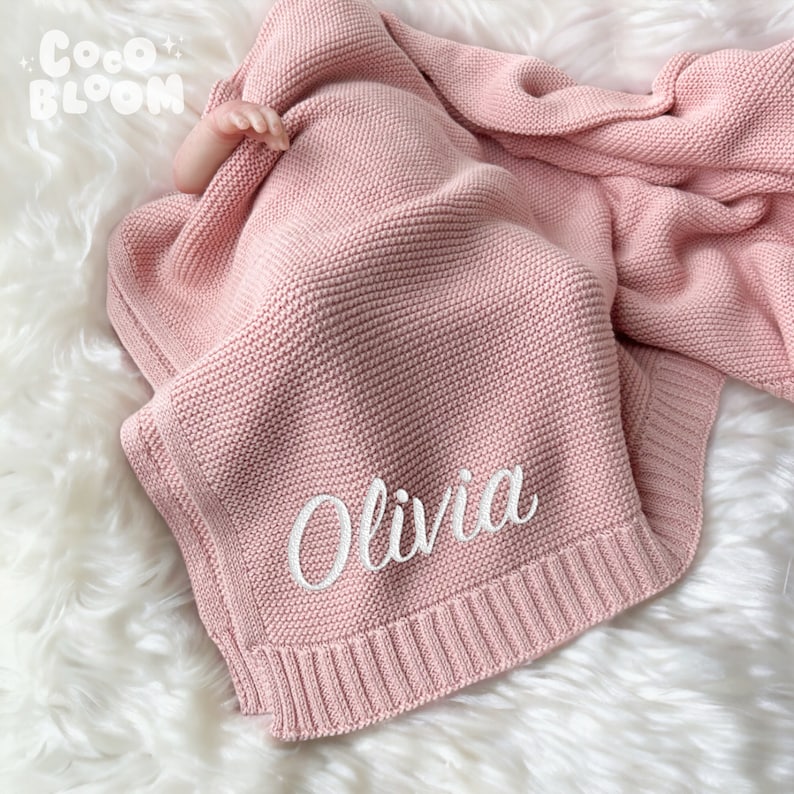 Op de afbeelding: Een zachtroze gebreide babydeken met de naam "Olivia" in het wit geborduurd. De deken is opgevouwen en gedrapeerd over een zacht, wit oppervlak. Het Coco Bloom-logo is zichtbaar.